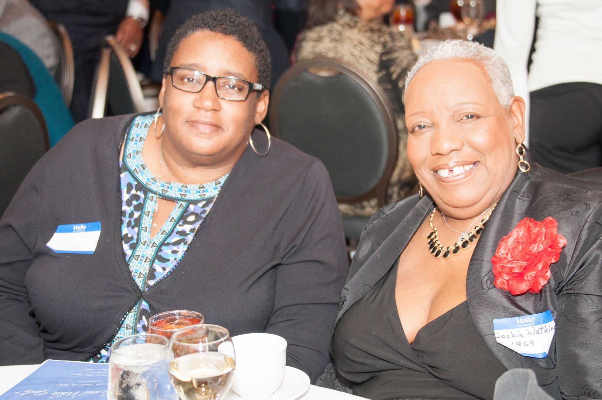 Two people seated at a table smiling. One wears glasses and a dark top. The other wears a black blazer with a flower.