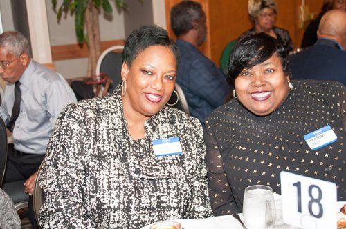 Two women smiling at a table; one in black and white jacket, the other in a black top with sequins. Table number 18.