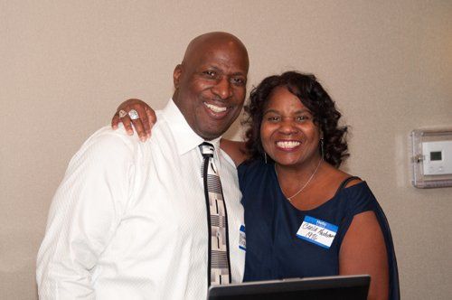 Two smiling people, man with arm around woman, posing indoors.