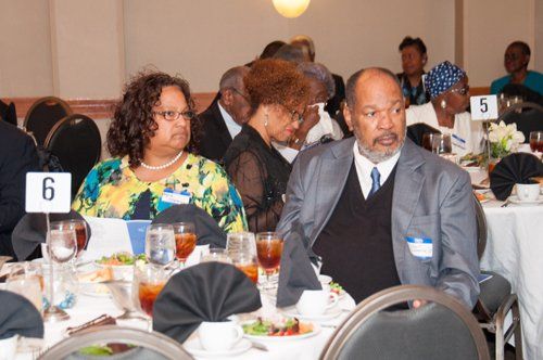 People seated at a formal banquet table. Woman in colorful blouse and man in gray blazer are in the foreground.