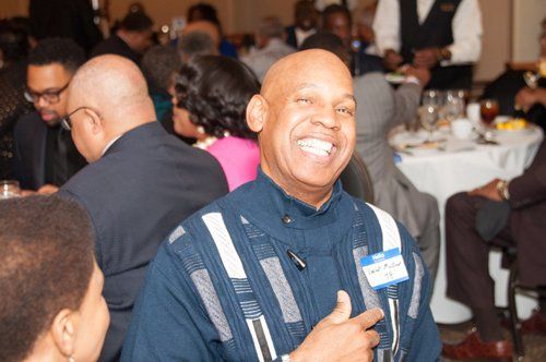 Smiling man in blue outfit at a banquet table, hand on chest, surrounded by others.