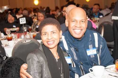 Smiling couple at a banquet table; woman in a silver jacket, man in a blue jacket. Others in background.