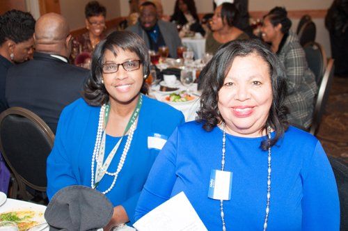 Two women in blue at a table, smiling at a conference. Others dine in the background.