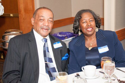 Two people at a table, smiling. Man in suit and tie. Woman in a blazer. At a dining event.