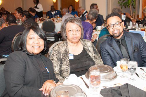 Three people seated at a table, smiling, in a well-lit banquet hall; others in the background.