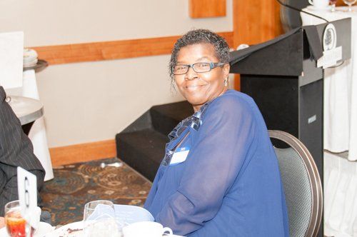 Woman in blue top, glasses, smiles at a table in a room with a podium.