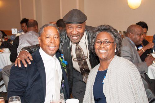Three people smiling together at a formal event, one has arm around another. Tables and other guests in background.