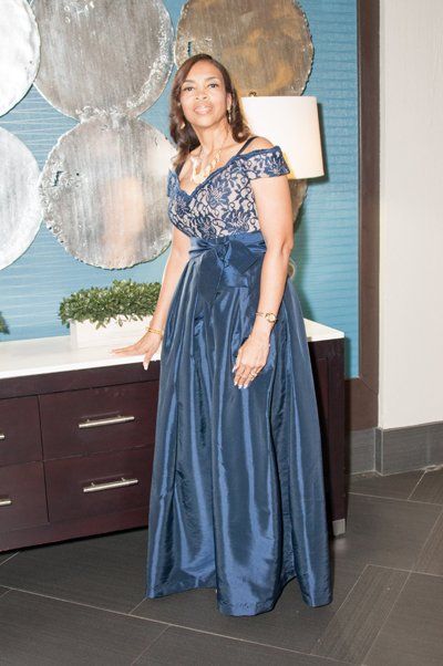 Woman in a navy blue formal dress, standing next to a table with a wall decoration in the background.