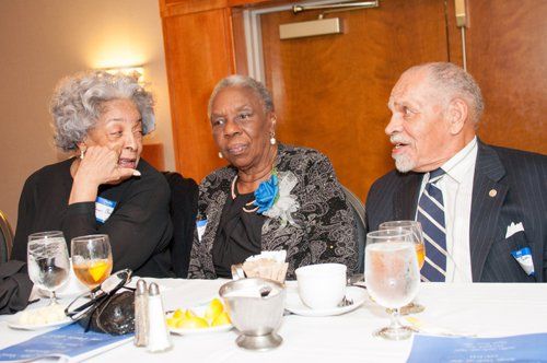 Three people at a table; two women, one man, conversing with drinks and food present.