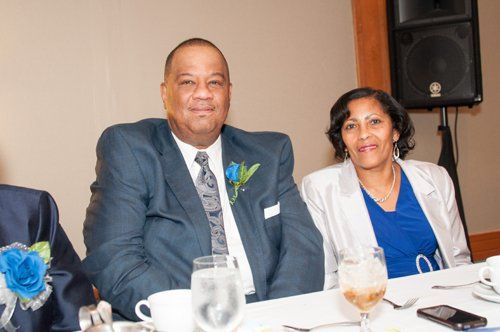 Man and woman seated at a table, both smiling, at an event. Man in blue suit, woman in white jacket.