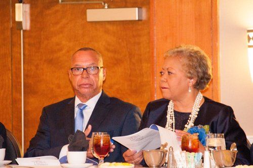Man and woman at table, man speaking, woman holding papers, formal setting.