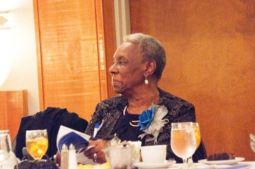 Woman at a table, wearing a dark jacket with a blue flower. She has short, grey hair and is looking to the side.