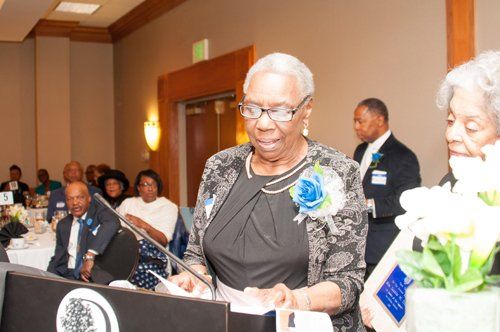 Woman at a podium speaking at a formal event, other attendees seated at tables.
