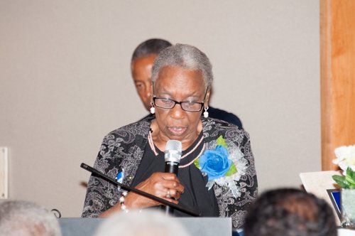 Woman speaking into a microphone at an event, wearing glasses and a floral pin. Another person stands behind her.