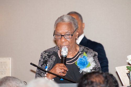 Woman with glasses speaking into a microphone; blue rose on her jacket.