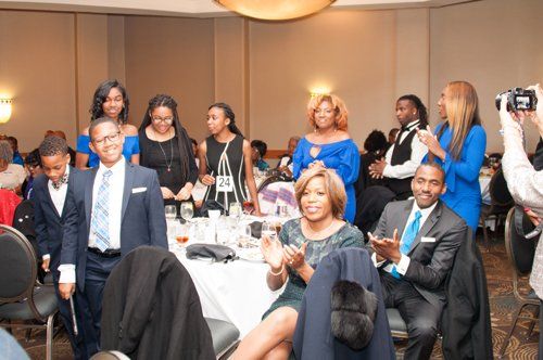 People gathered at a formal event, some clapping. Several people wear blue. Hotel ballroom setting.