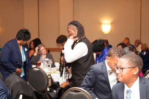 People at a formal event, some standing, some seated at tables.  Man in vest talking. Others in suits and dresses.