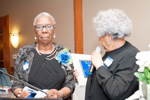 Two women at an event: one receiving a plaque, the other speaking into a microphone.
