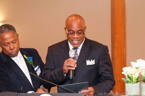 Man in suit speaks into microphone at an event, another man looks on.