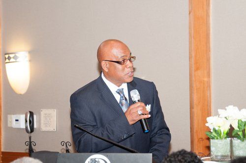 Man in a suit speaks into a microphone at a podium. Neutral background with a lamp and flowers.