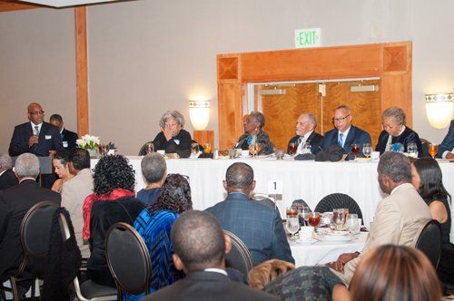 A panel of people sits at a table while a man speaks at a formal event.