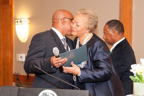 Man kisses woman on the cheek, holding a microphone and document at an event. Another man looks on.
