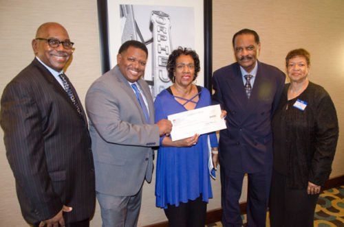 Five people in suits, one woman receiving a check; they are smiling in an indoor setting.