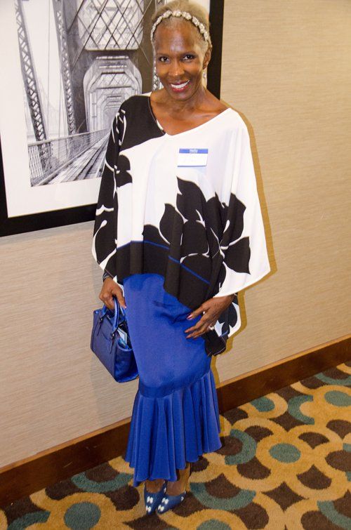 Woman in black and white top and blue skirt, holding a blue bag, standing in front of a framed bridge photo.