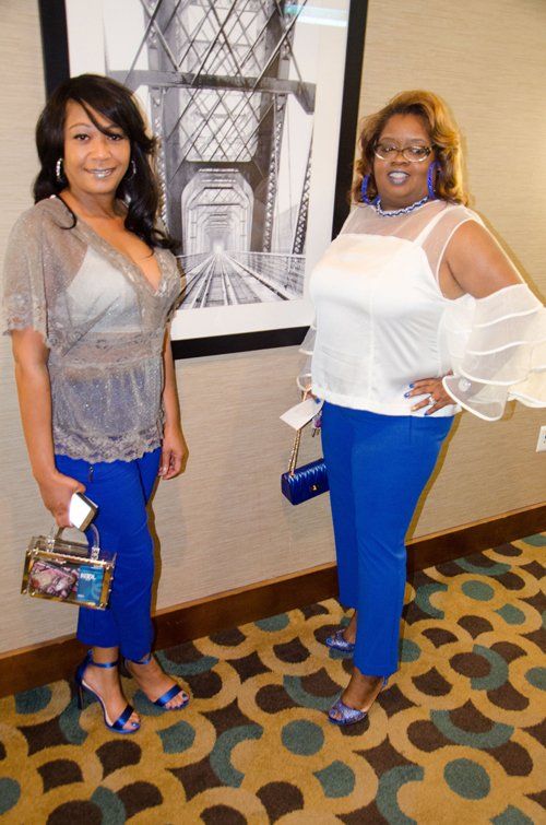 Two women pose, both in blue pants, different tops. One holds a purse. Black and white artwork behind them.