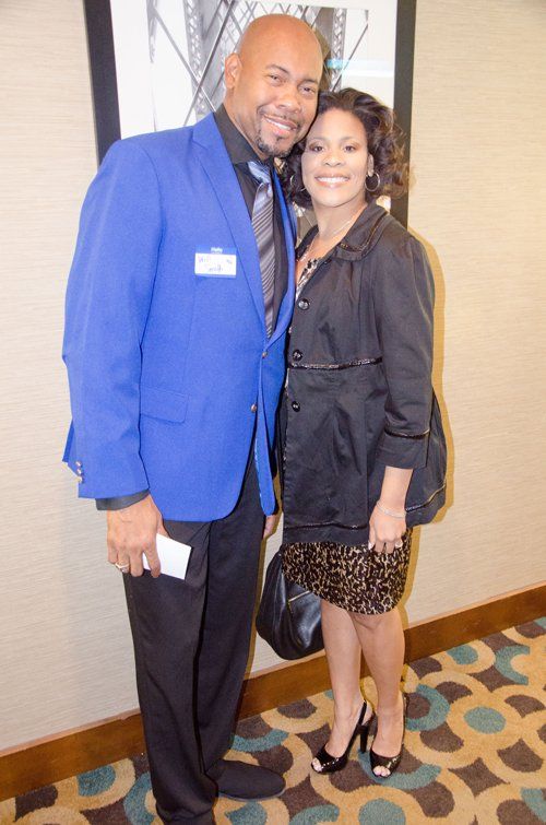 Man in blue blazer and woman in black jacket pose together, smiling. Formal setting.