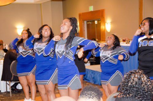 Cheerleaders in blue and gray uniforms performing at an event, arms raised, smiles.