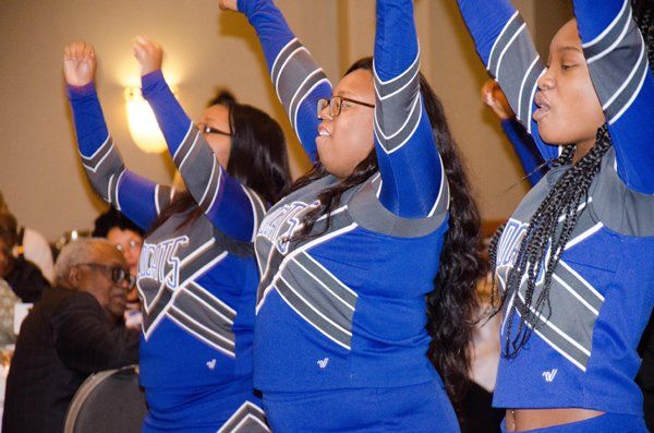 Cheerleaders in blue and gray uniforms with arms raised, performing at an event.