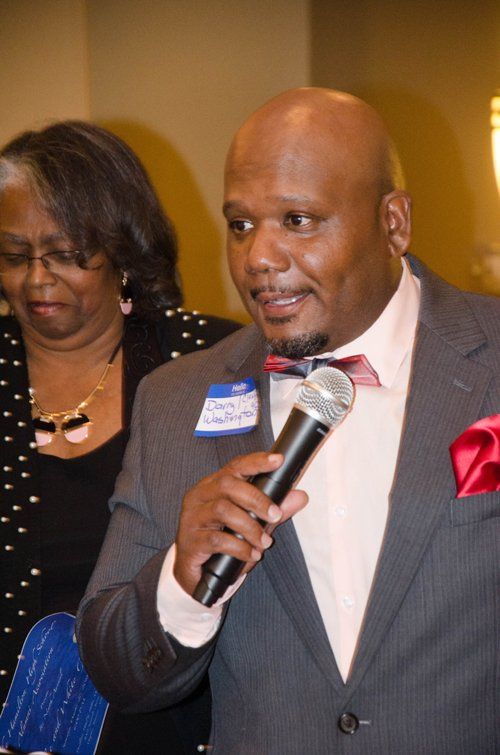 Man in suit speaking into a microphone; woman in background.