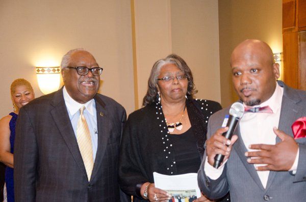 Man speaking into a microphone, flanked by a man in a suit and a woman. All are indoors.