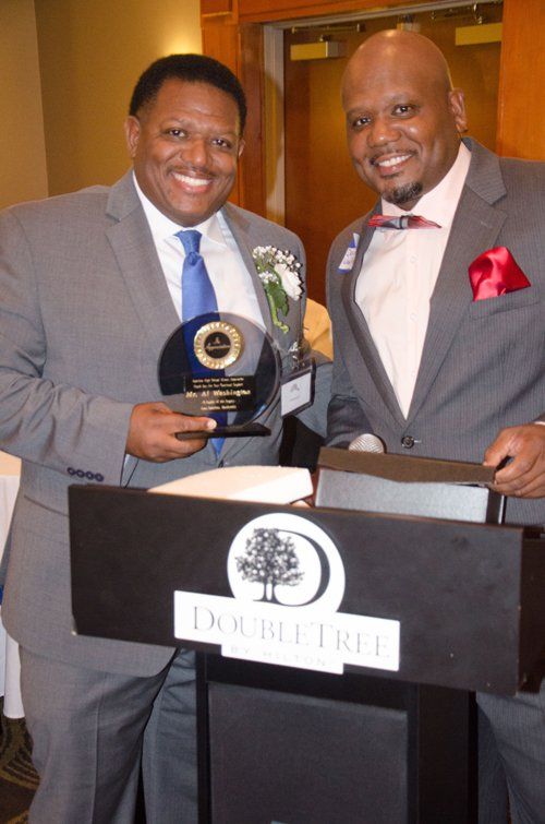 Two men in suits at a podium; one holds an award. They are smiling, with a 