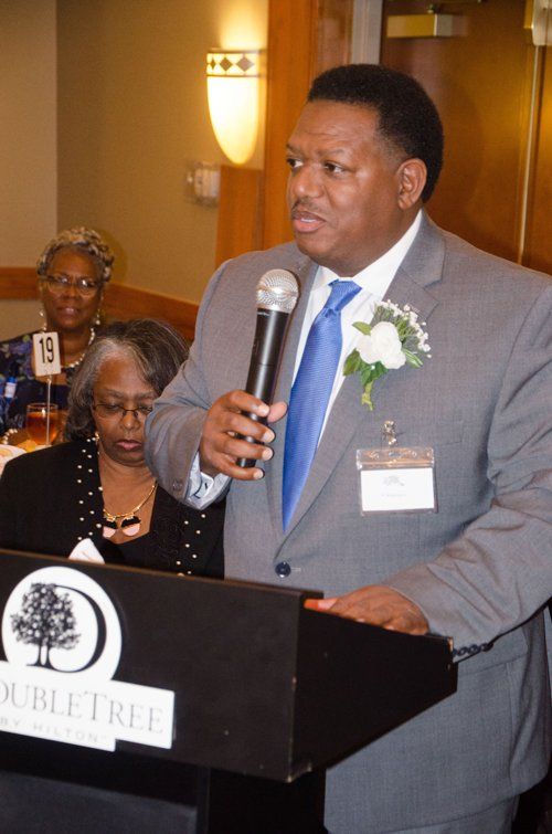 Man in grey suit speaking into a microphone at a DoubleTree event; two women in the background.