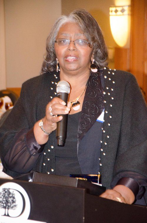 Woman speaking into a microphone at a podium, wearing a black jacket with white polka dots, and holding a microphone.