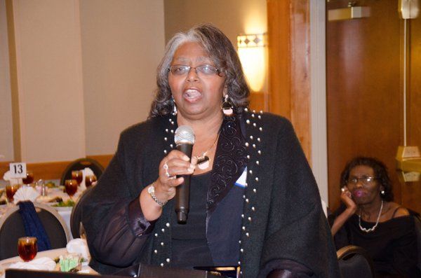 Woman speaking into a microphone at an event, wearing a black blazer.