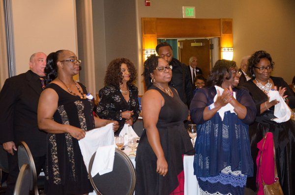 People standing at a banquet table; some holding napkins, clapping, and looking towards an event.