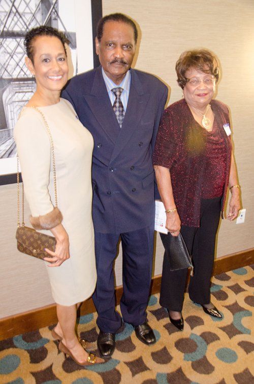 Three people pose for a photo in a carpeted room. The man in a dark suit stands between two women.