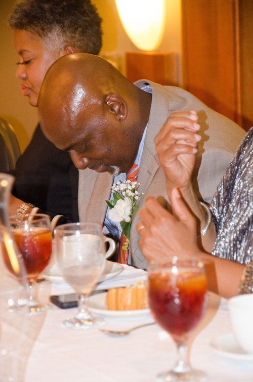Man in suit bows head at a table setting, drinks and other people partially visible.