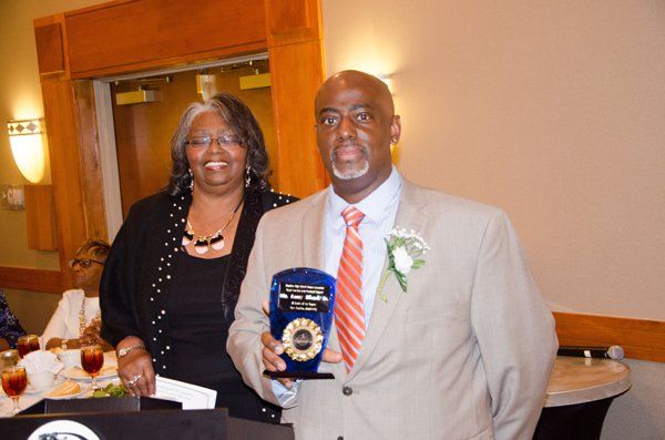 Man in suit holds award, woman smiles next to him. Formal event, beige walls.