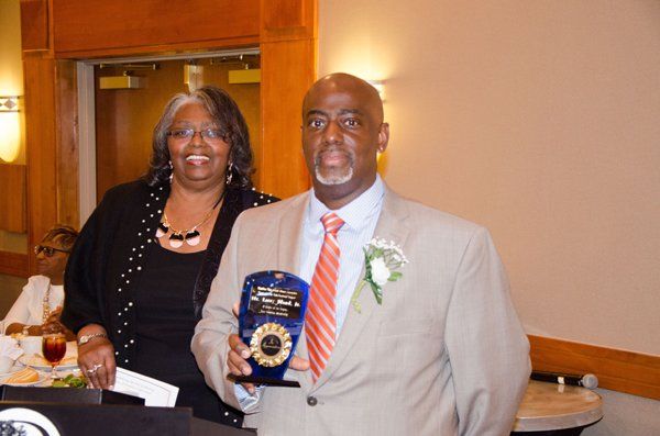Man in a suit holding an award, woman beside him smiling. Indoors, warm lighting.