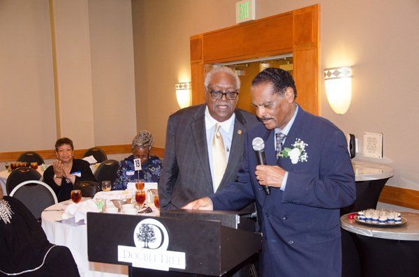 Two men at a podium; one speaks into a microphone, the other looks on. Indoors, at an event.