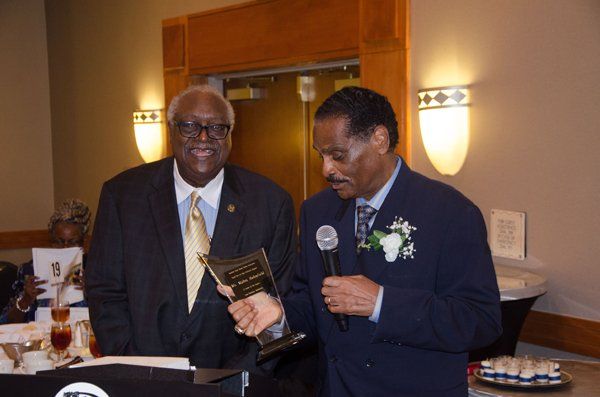 Two men at an event, one holding an award and a microphone, smiling, formal setting.