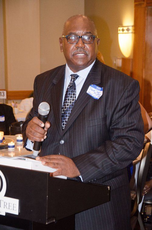 Man in suit speaking at a podium, holding a microphone. Dimly lit room, possibly at a formal event.