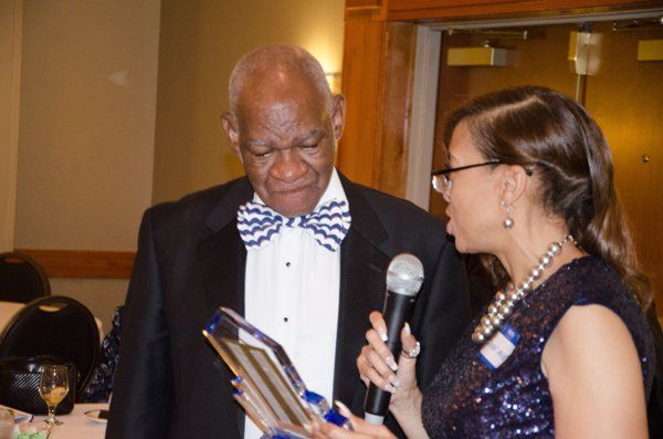 Woman speaking into microphone, presenting an award to a man in a formal setting.