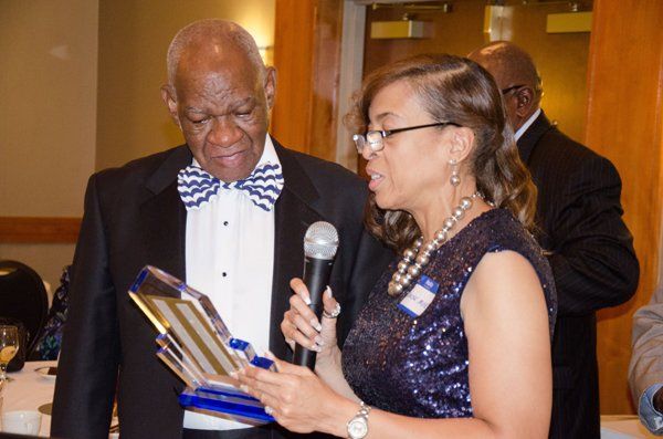 Woman presenting an award to a man in a formal setting. The woman holds a microphone and the award.