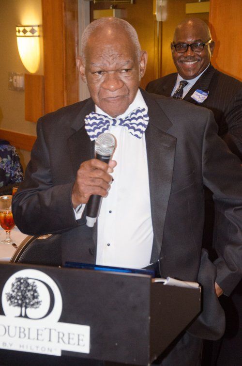 Man speaking into a microphone at a DoubleTree event, wearing a bow tie and suit. Another man smiles in the background.