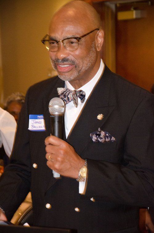 Man in a black blazer speaks into a microphone, wearing glasses and a patterned bow tie, in a lit room.